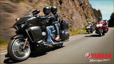 two people riding on the back of a black motorcycle down a mountain road with mountains in the background