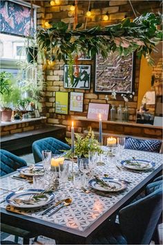 a dining room table with place settings and candles on it, surrounded by greenery