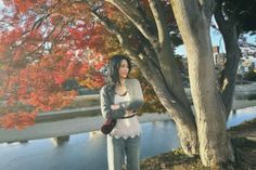 a woman standing next to a tree in front of a lake with red leaves on it