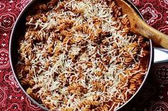 a skillet filled with pasta and cheese on top of a red table cloth next to a wooden spoon