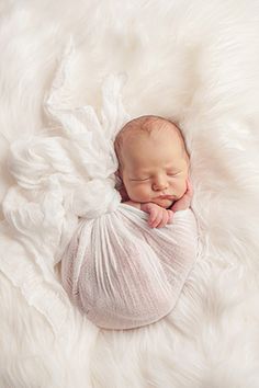 a newborn baby wrapped in a white wrap is laying on a fluffy white blanket with his hands under his chin