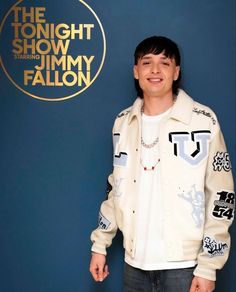 a young man standing in front of a blue wall wearing a white jacket and jeans