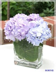 there is a small vase with purple flowers in it on the table near some bushes