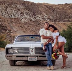 a man and woman standing next to a car in front of a mountain side area