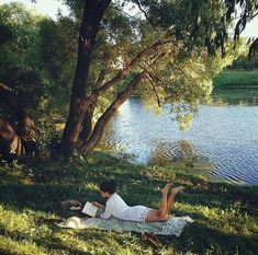 a woman laying on the grass next to a lake reading a book while sitting under a tree