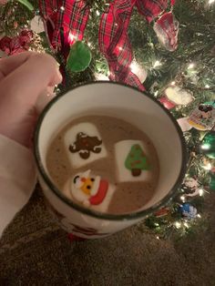 someone holding a cup of hot chocolate with marshmallows in it and christmas decorations on the tree