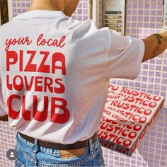 a man holding a pizza box in his right hand and wearing a t - shirt that says your local pizza lover's club