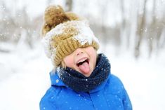 a young boy wearing a blue coat and hat in the snow with his tongue out