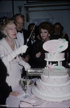 an older woman cutting into a wedding cake with her family and friends in the background