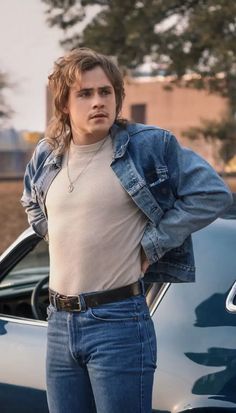 a young man standing in front of a blue car wearing a jean jacket and jeans