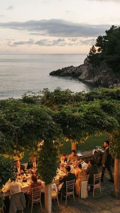 a group of people sitting around a dinner table near the ocean with trees and bushes