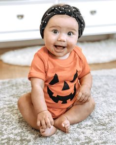 a baby sitting on the floor wearing a pumpkin shirt