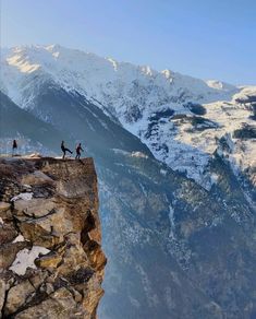 two people standing on the edge of a cliff with snow covered mountains in the background