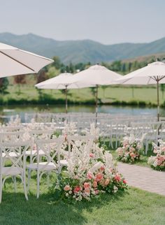 an outdoor wedding set up with white chairs and flowers on the lawn, surrounded by umbrellas