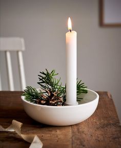 a white candle is sitting in a bowl with pine cones and greenery on the table