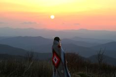 a person standing on top of a hill with a blanket over their shoulders and mountains in the background