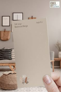 a person holding up a beige paint swatch in front of a room with pictures on the wall