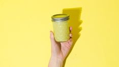 a hand holding a glass jar filled with green liquid against a yellow background that is half empty