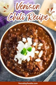 a white bowl filled with chili and onions next to a cutting board, spoons and utensils