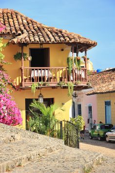 a yellow house with pink flowers on the balcony and steps leading up to it's second story