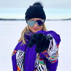 a woman wearing sunglasses and a purple jacket pointing at the camera with her hand in the snow