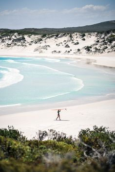 a person with a surfboard walking on the beach near some blue water and white sand