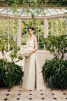 a woman in a white dress holding a bouquet and standing next to some potted plants