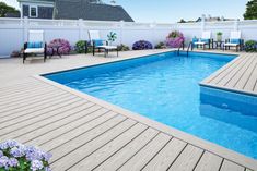 a backyard with a pool and decking surrounded by purple hydrangeas, blue chairs and white picket fence