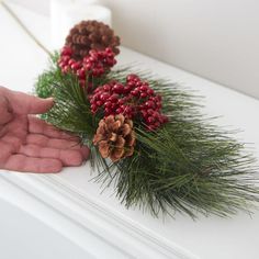 a hand is holding pine cones and red berries on a white mantle with greenery