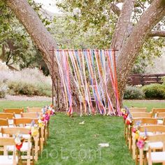 an outdoor ceremony set up with wooden chairs and colorful streamers hanging from the trees