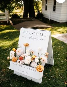 a welcome sign with flowers on it in front of a white house and grass area