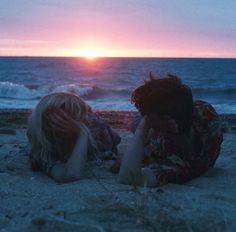 two people laying on the beach with their heads in their hands as the sun sets