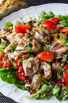 a salad with chicken, tomatoes and lettuce on a white plate next to a piece of bread