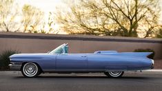 an old blue convertible car parked in front of a building with trees and bushes behind it