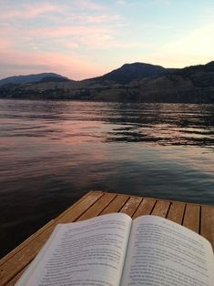 an open book sitting on top of a wooden dock next to the water with mountains in the background