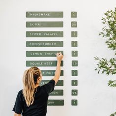 a woman writing on a wall with green street signs in front of her and a potted plant next to it