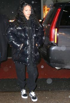 a woman in black jacket and sneakers standing next to a car with snow falling on it