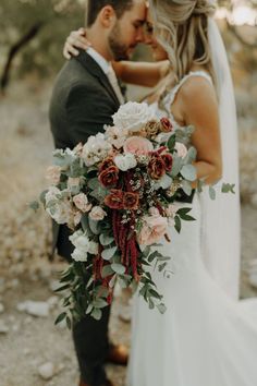 a bride and groom standing together in the woods with their arms around each other holding a bouquet