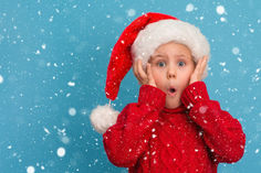 a young boy wearing a santa hat covering his face with both hands while standing in the snow