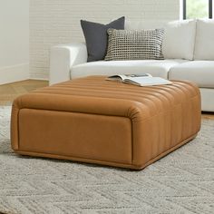 a tan ottoman sitting on top of a rug in front of a white couch and window