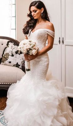 a woman in a wedding dress holding a bouquet and looking at the camera while standing next to a couch