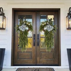 two wreaths on the front door of a white house with black planters and lanterns