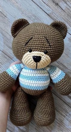 a crocheted brown teddy bear sitting on top of a wooden floor next to a person's hand