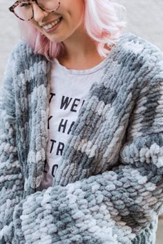 a woman with pink hair and glasses is wearing a gray cardigan sweater over a white t - shirt