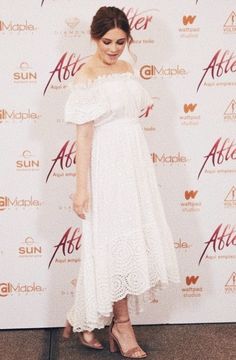 a woman standing in front of a wall wearing a white dress and high heeled sandals