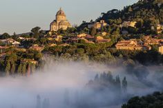 fog covering the ground in front of a small town on top of a hill with trees