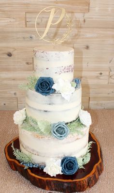 a three tiered wedding cake with blue flowers and greenery sits on a wooden platter