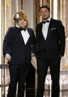 two men in tuxedos standing next to each other at an awards ceremony, one with a teddy bear on his head