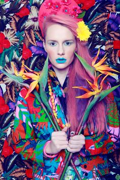 a woman with pink hair and flowers on her head is standing in front of a colorful background