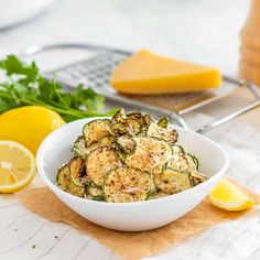 a white bowl filled with zucchini and parsley next to lemon wedges